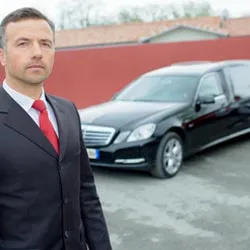 Chauffeur de pompes funèbres en costume sombre posant devant un corbillard noir Mercedes